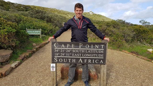 A man standing behind the Cape Point sign with its coordinates.