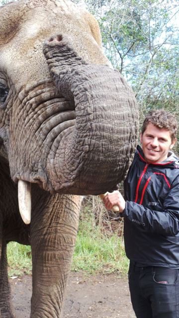 A man smiling next to an elephant's head and curled trunk.