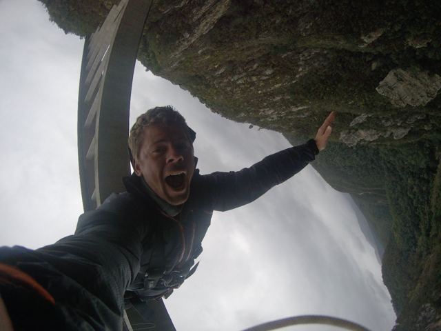 A man screaming while hanging upside down on a bungee cord beneath a bridge.