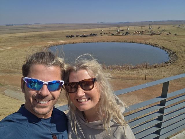 A man and a woman in sunglasses taking a selfie above a round waterhole on flat grassland.