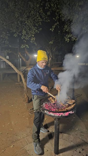 A man in a beanie grilling meat on a smoking braai at night.