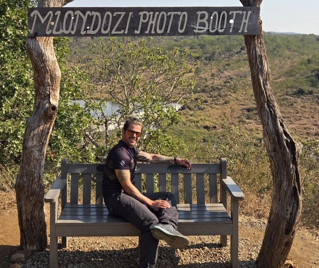 Man sitting on a wooden bench under a rustic Mlondozi Photo Booth sign overlooking the bush.