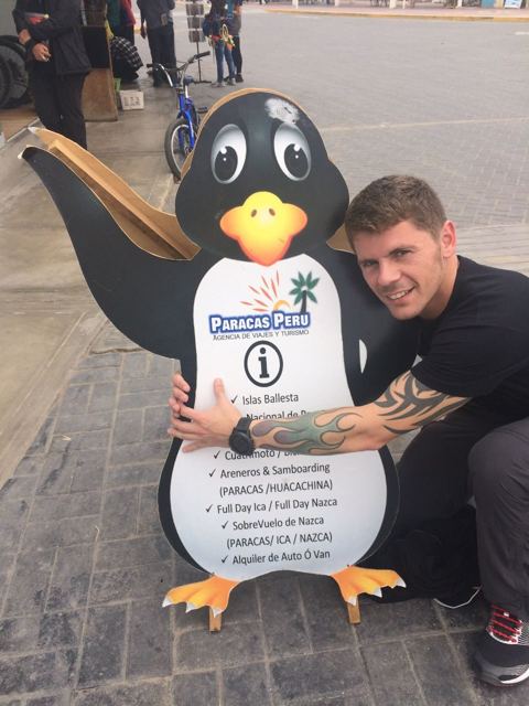 A man crouching next to a cartoon penguin information sign in Paracas.