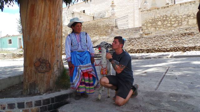 A man kneeling beside a llama next to a woman in traditional Andean dress in a village square.