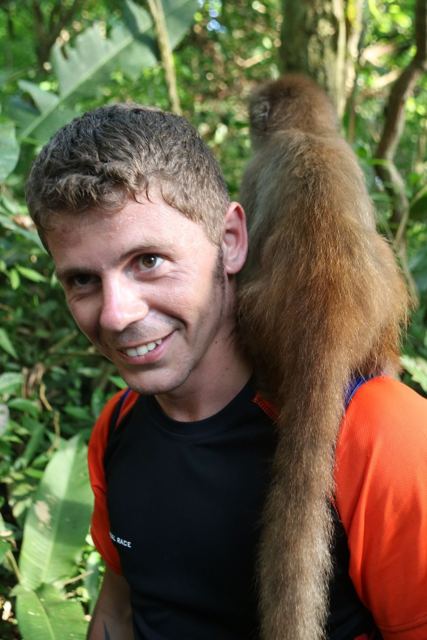 A smiling man with a brown monkey perched on his shoulder among jungle leaves.
