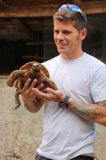 A smiling man holding a small tortoise at an animal sanctuary.