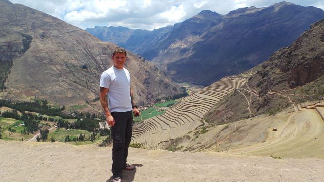 A man standing on a hillside above terraced fields and mountains in the Sacred Valley.