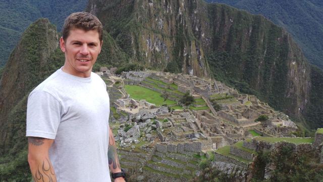A man in a white T-shirt in front of the ruins of Machu Picchu and Huayna Picchu.