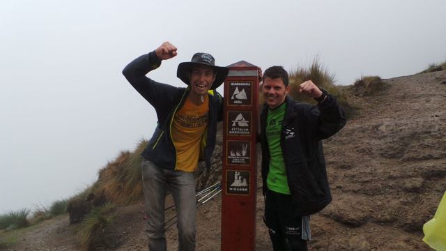 Two hikers flexing beside a trail marker on a misty mountaintop near Machu Picchu.