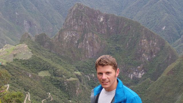 A man in a blue jacket with the steep green peak of Huayna Picchu behind him.