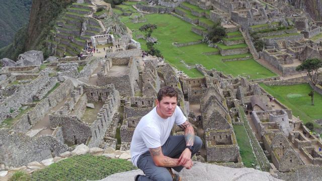 A man crouching on Inca stone walls with the terraces of Machu Picchu behind.