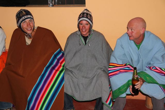 Three men wrapped in colourful Andean ponchos and woolly hats sitting in a room.