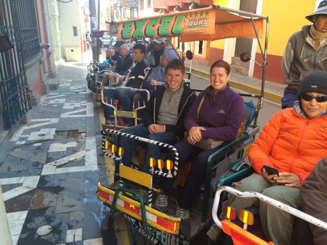 A group of travellers sitting in an open-sided tourist vehicle on a colourful street in La Paz.