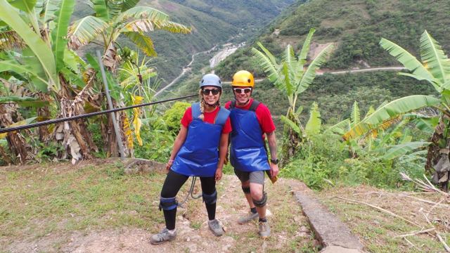 Two people in harnesses and helmets standing on a jungle hillside before a zipline.