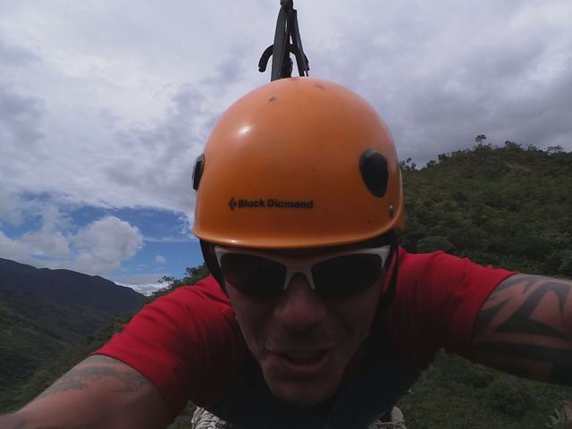 A man in an orange helmet and sunglasses taking a selfie while hanging from a zipline.
