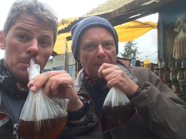 Two men drinking from plastic bags of tea through straws at a roadside stall.