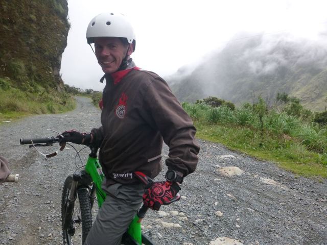 A man in a helmet and riding gear standing with a mountain bike on a gravel mountain road.