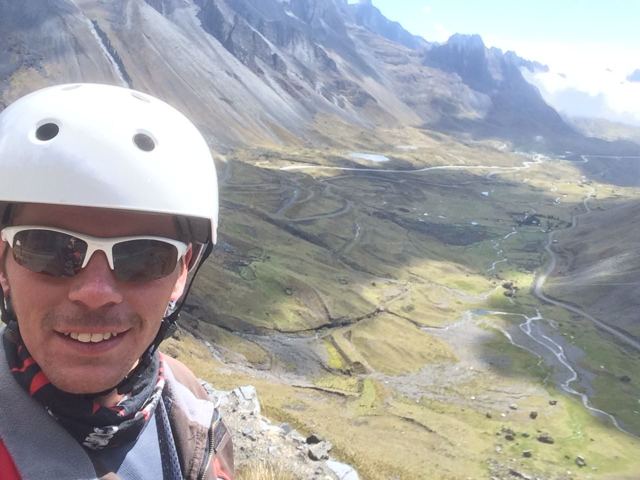A smiling man in a white helmet and sunglasses above a green valley with a winding road.