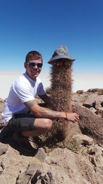 A man crouching beside a tall cactus wearing a hat and sunglasses, with the white salt flats behind.