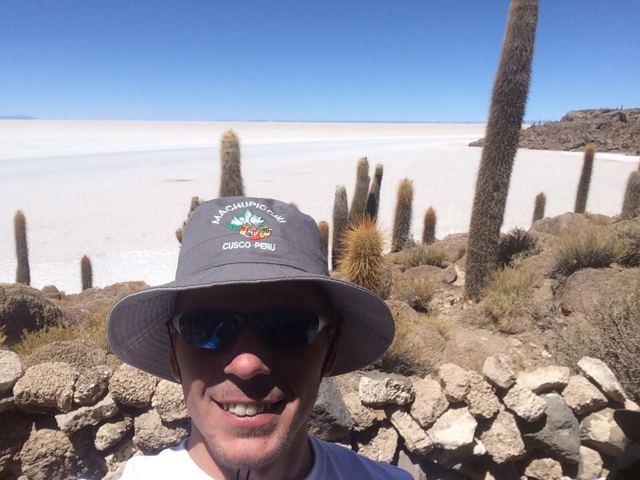 A man in a sun hat taking a selfie among giant cacti with the salt flats stretching behind.