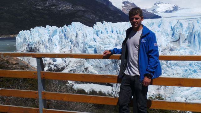 A man leaning on a railing with the huge ice face of Perito Moreno Glacier behind him.