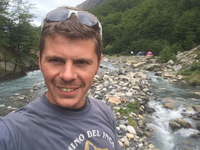A smiling man with sunglasses on his head beside a rocky glacial river in Patagonia.