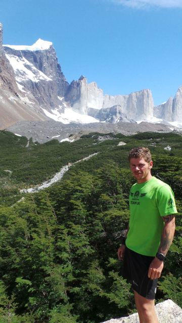 A man in a green T-shirt on a trail with the granite spires of Torres del Paine behind.