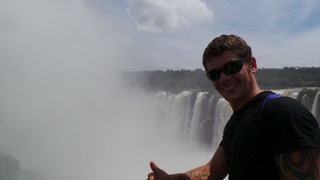 A man in sunglasses giving a thumbs up in front of misty Iguazú Falls.