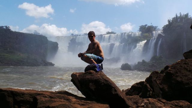 A man crouching on a rock with the wide curtain of Iguazú Falls behind him.