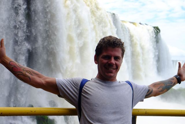 A man with arms spread wide in front of thundering white waterfalls at Iguazú.