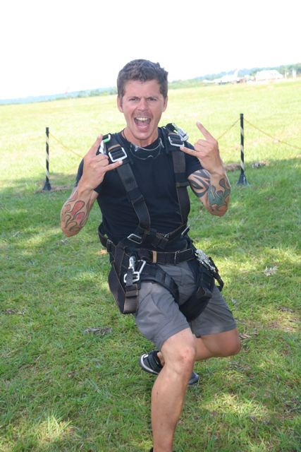 A man in a skydiving harness making rock-on hand signs on a grassy landing field.