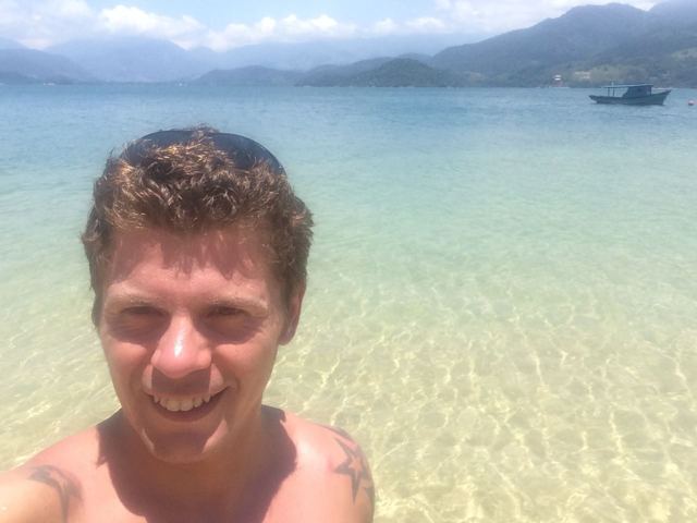 A smiling man taking a selfie in crystal-clear shallow sea water with green hills behind.