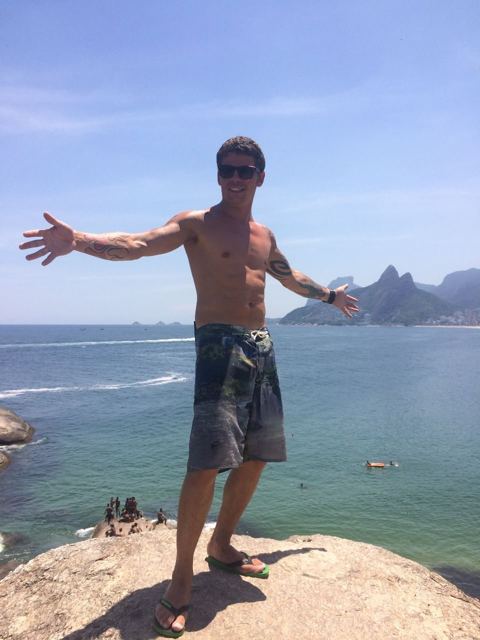 A man in board shorts standing with arms out on a rock above the sea in Rio.