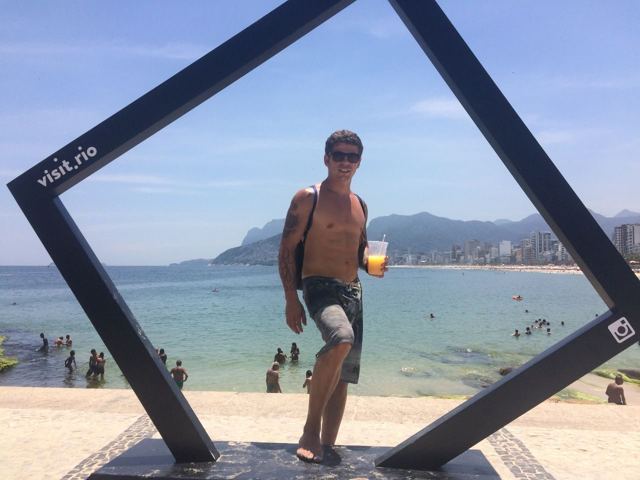 A man with a drink posing inside a large photo frame sculpture on Ipanema beach.