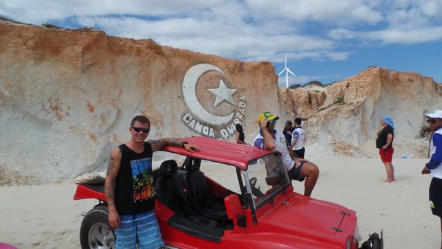 A man standing by a red beach buggy beneath sandstone cliffs with a carved moon and star.