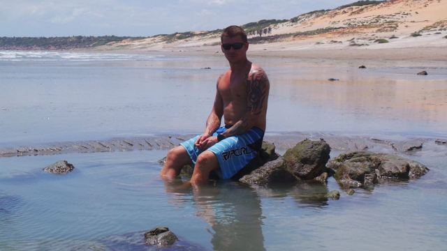 A man sitting on rocks in shallow water on a wide beach with dunes behind.