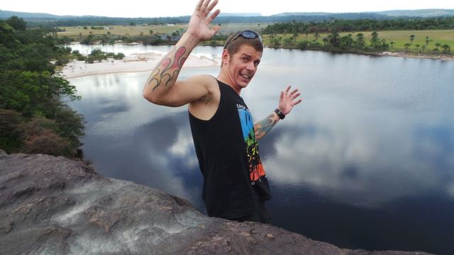 A man waving on a rock above a calm lagoon with forest on the far shore.