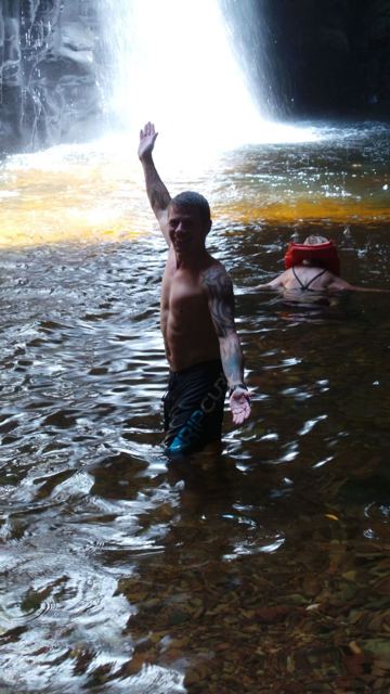 A man raising one arm while standing in water beneath a waterfall.