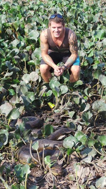 A man crouching among leaves with a coiled anaconda in the foreground.