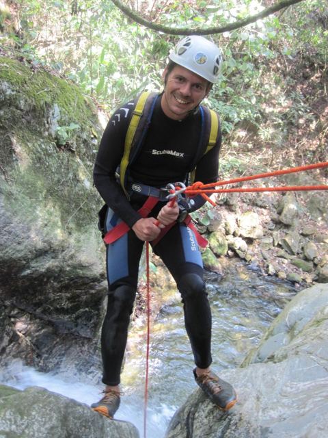 A man in a wetsuit and helmet abseiling down a rock face beside a waterfall.
