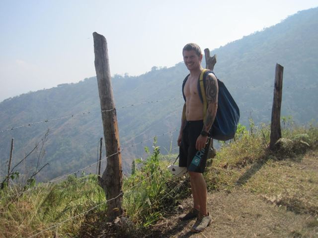A man with a rucksack on a grassy ridge between fence posts above a valley.