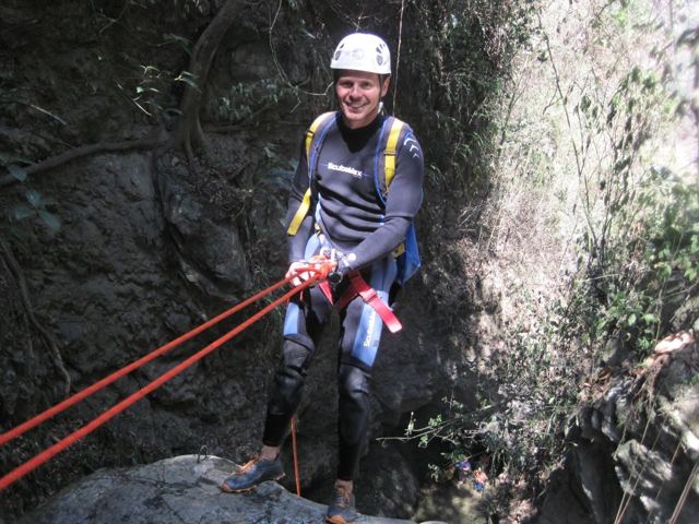 A man in a wetsuit and helmet abseiling into a mossy gorge.