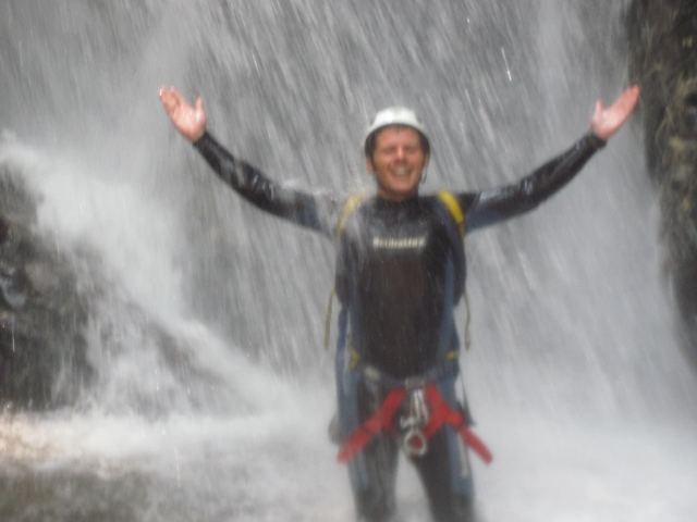A man in a wetsuit and helmet with arms out under a pounding waterfall.