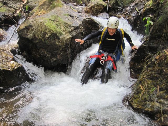 A man in a wetsuit and helmet sliding down a rocky waterfall chute.