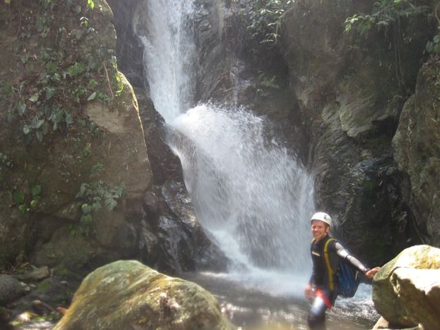 A man in a helmet standing on rocks at the foot of a tall waterfall in a gorge.
