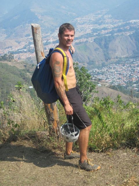A man with a rucksack holding a helmet on a hillside above the city of Mérida.