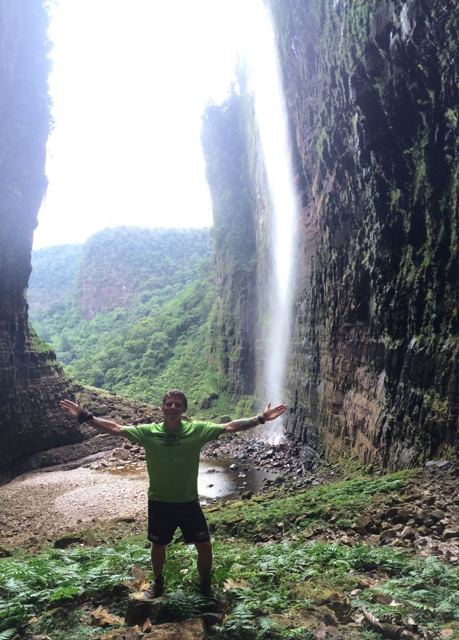 A man with arms outstretched at the bottom of a tall waterfall between towering cliffs.