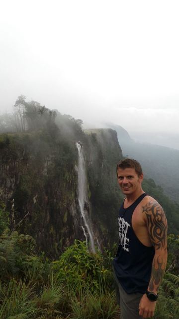 A man in the foreground with the misty cascade of Angel Falls dropping from a cliff behind.