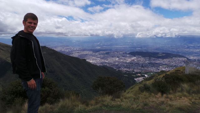 A man in a dark jacket on a mountainside with the city of Quito spread out below.