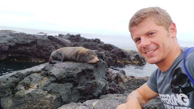A smiling man taking a selfie with a sea lion lying on the rocks behind him.